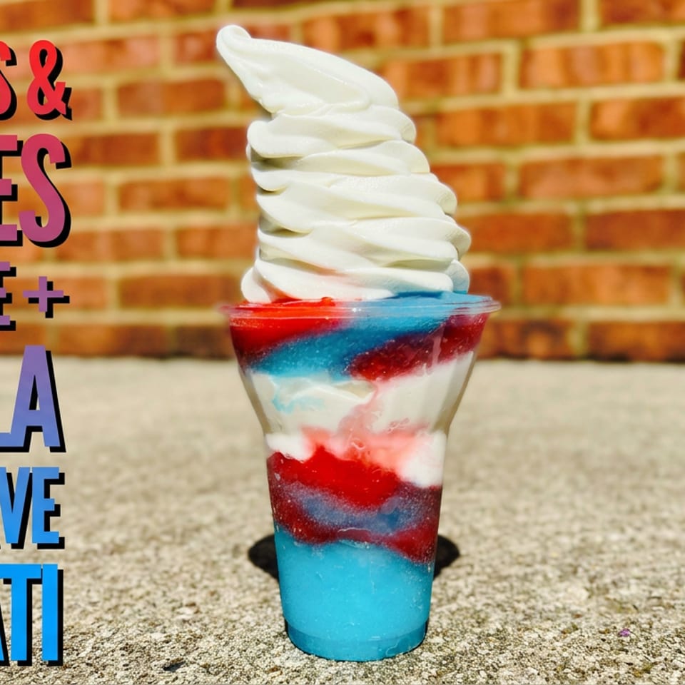 Layered red, white, and blue Italian ice topped with vanilla soft serve in a cup against a brick wall.