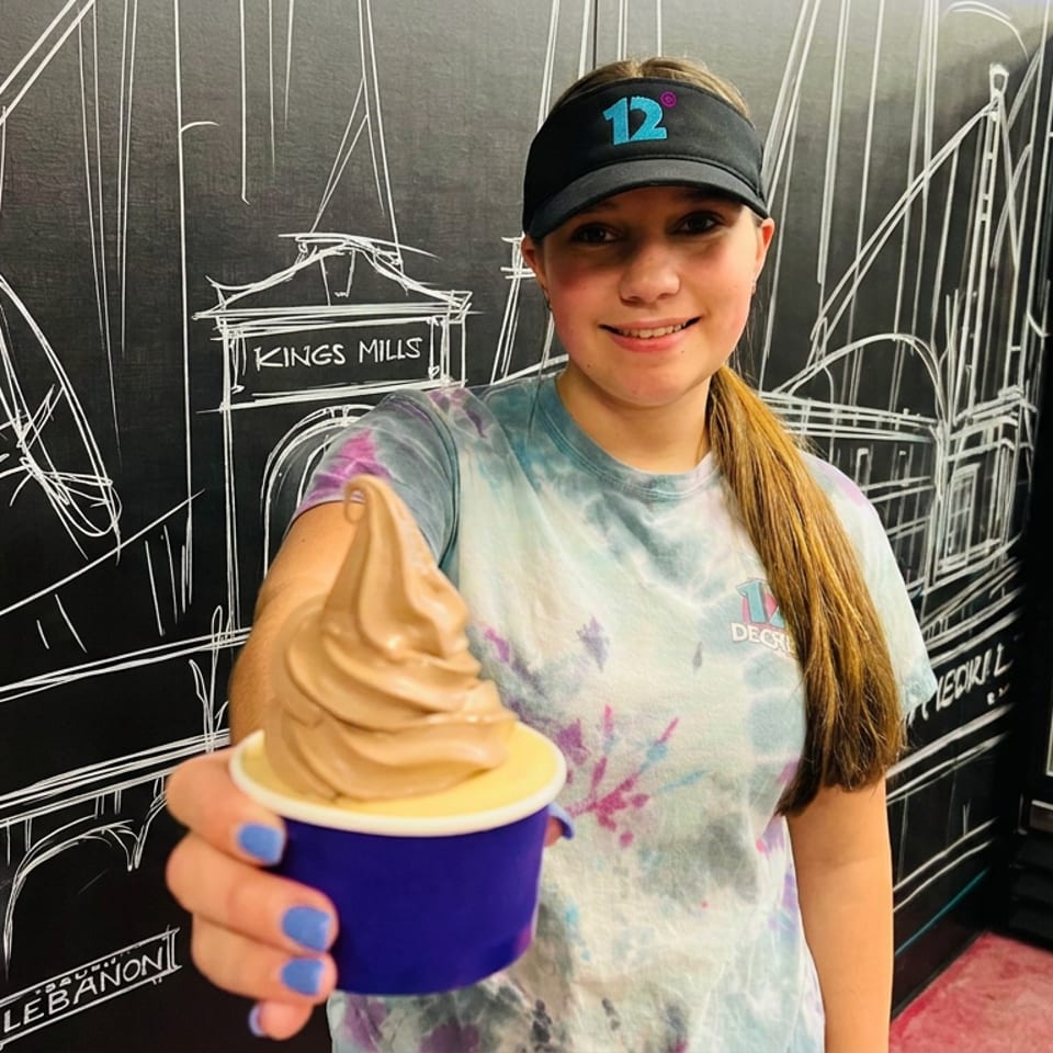 Smiling restaurant worker holds a cup of soft-serve ice cream in front of a cityscape mural and beverage refrigerator.