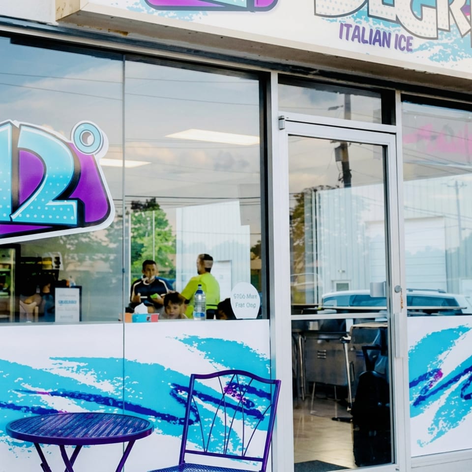 Colorful L2 Italian Ice storefront with wave graphics, outdoor seating, glass doors, and customers visible inside.