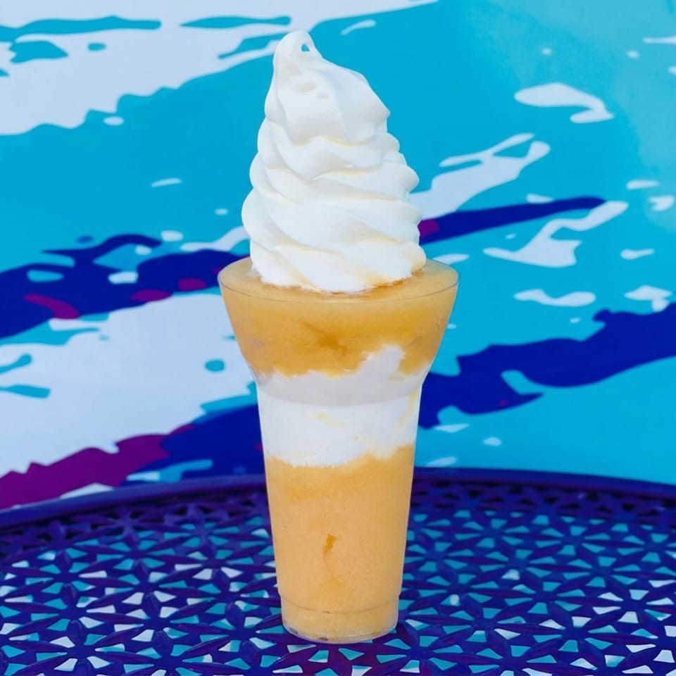 Layered golden dessert drink topped with a tall swirl of vanilla soft serve on a colorful outdoor table.